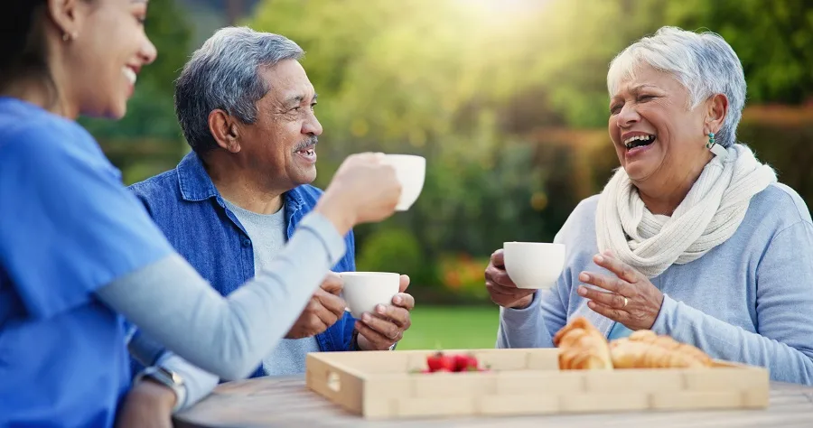 elderly couple having tea time outdoors with their 24/7 caregiver who is a young woman